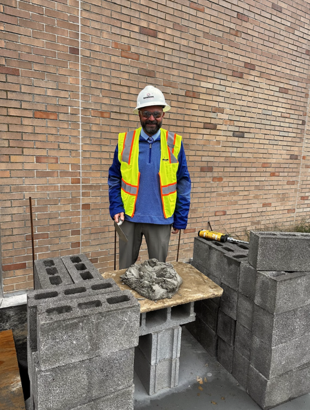 A man in a hard hat and yellow safety vest prepares a brick for placement on a construction site.