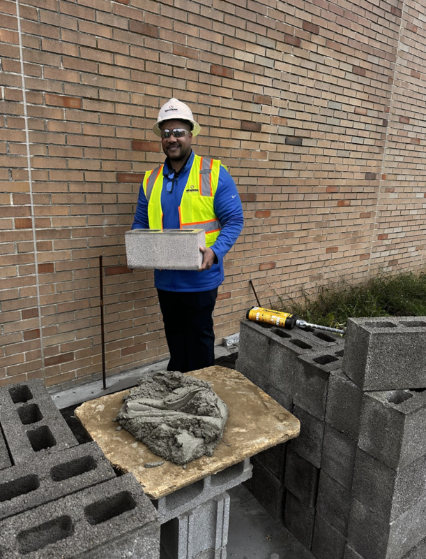 A man in a hard hat and yellow safety vest prepares a brick for placement on a construction site.