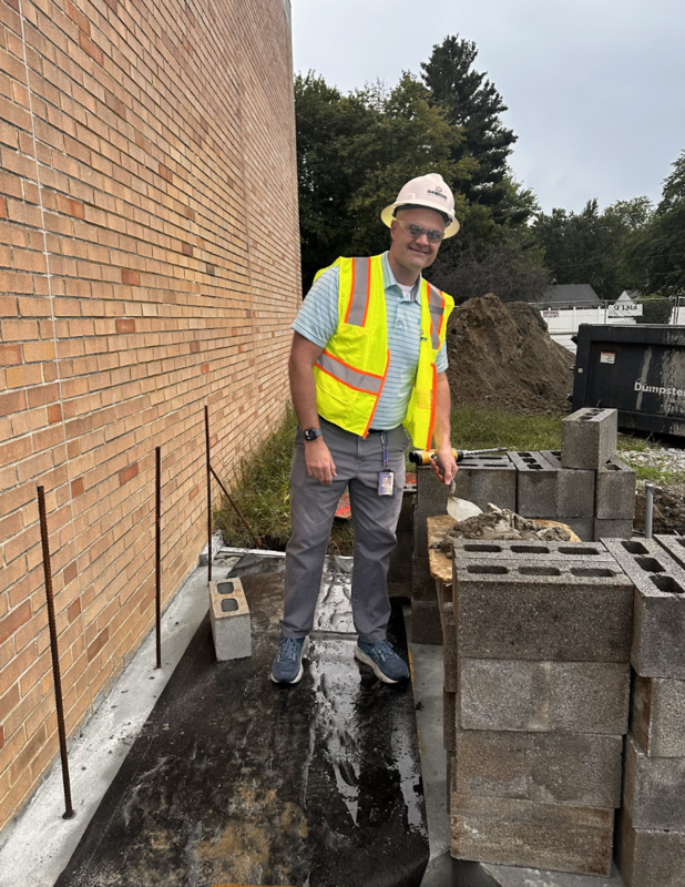 A man in a hard hat and yellow safety vest prepares a brick for placement on a construction site.