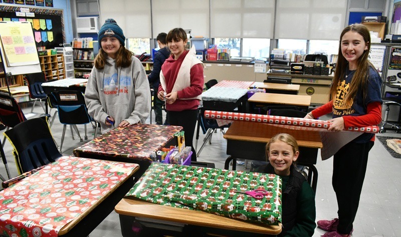 smiling students decorate classroom desks with festive holiday wrapping paper.