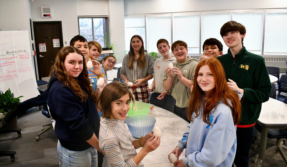A group of smiling young people gathers around a table in a bright room, holding bowls
