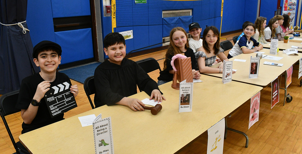 Students showcase their projects at a school science fair, featuring creative displays on various topics, including filmmaking and hobbies, in a lively gymnasium setting.