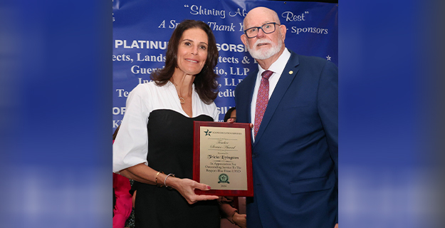 A woman in a white and black dress holds a plaque alongside a man in a blue suit with a red tie. They stand smiling at an award ceremony.