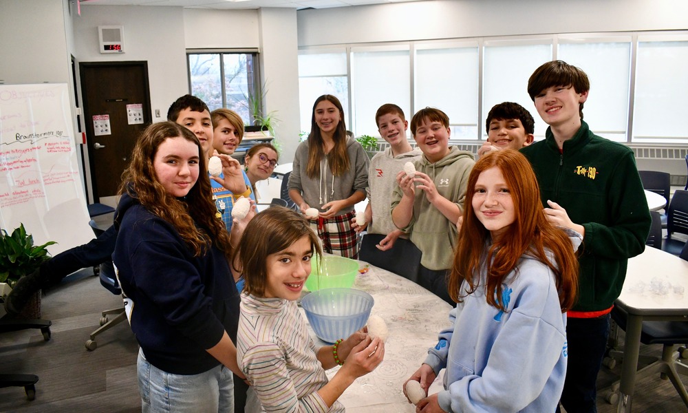 A group of smiling young teenagers stands around a table in a bright classroom. 