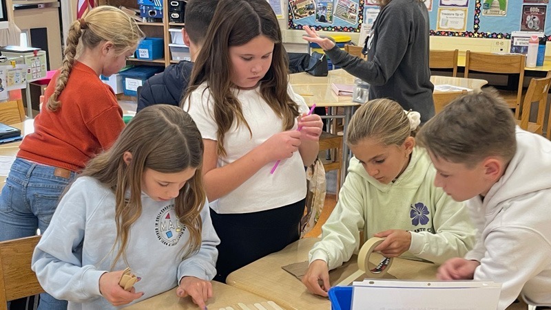 Children engaged in a classroom activity, working collaboratively with craft materials like popsicle sticks and masking tape. The atmosphere is focused and creative.