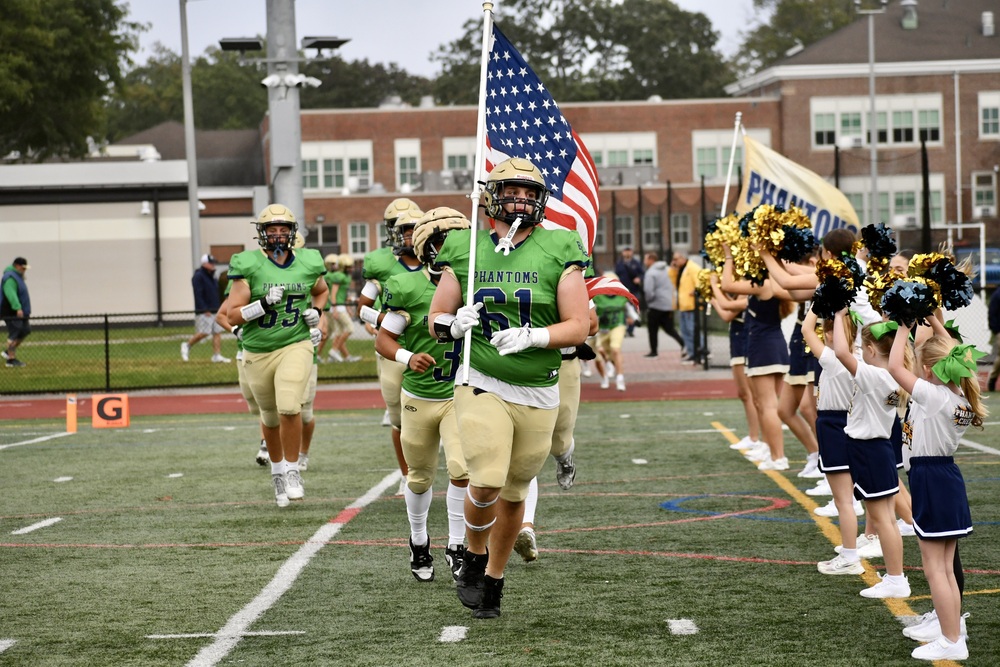 Student football players running flag, with cheerleaders standing beside them.