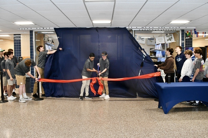 Students participate in a ribbon-cutting ceremony at school, unveiling a new space, with a blue curtain in the background and a table set up nearby.