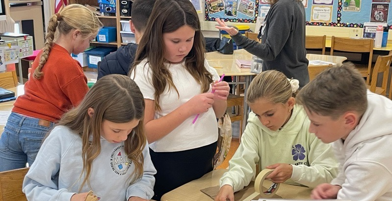 Children engaged in a classroom activity, working collaboratively with craft materials like popsicle sticks and masking tape. The atmosphere is focused and creative.