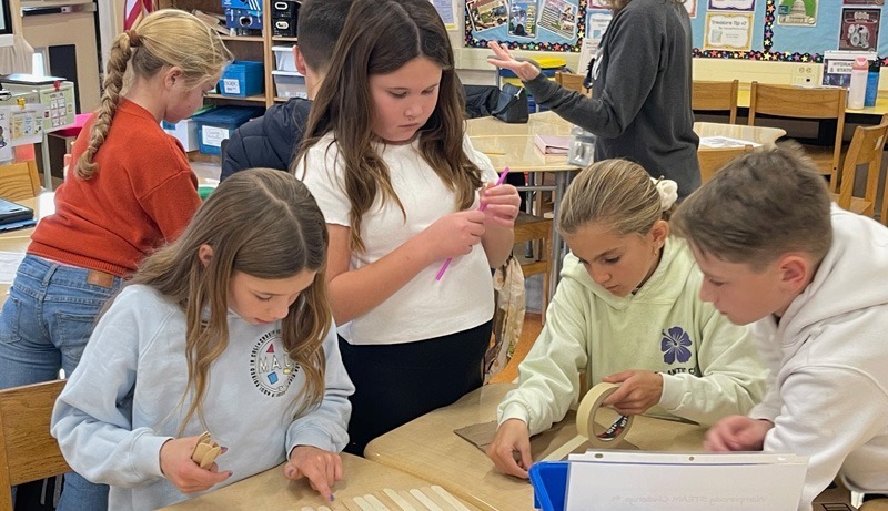 Children engaged in a classroom activity, working collaboratively with craft materials like popsicle sticks and masking tape. The atmosphere is focused and creative.