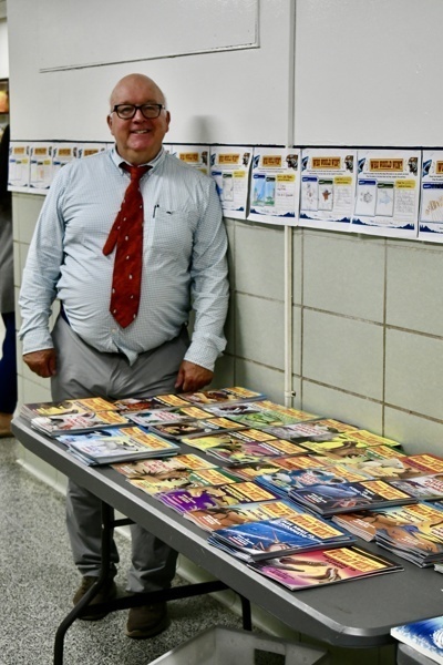 A person stands beside a table displaying various magazines in a classroom setting.