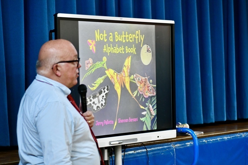 A person stands beside a screen displaying the cover of Not a Butterfly Alphabet Book.
