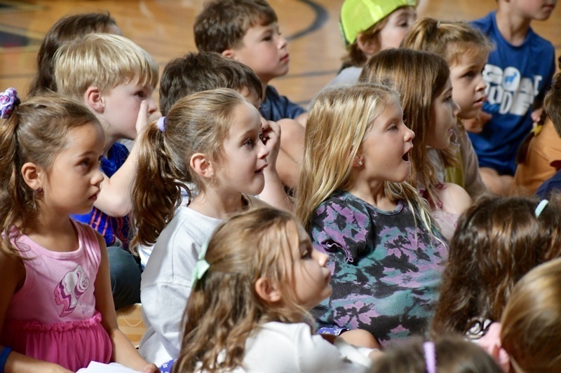 A group of children attentively watch an event in a gymnasium setting.