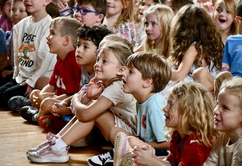A group of children sitting on the floor, attentively watching an event or performance.