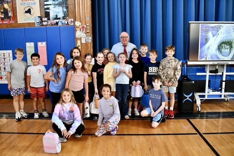A group poses together in a gymnasium with blue curtains and a presentation screen.