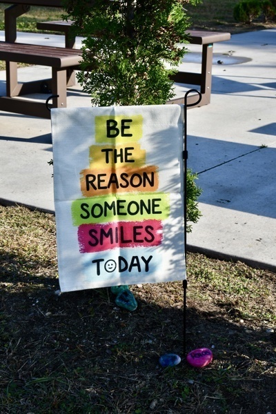 A sign with colorful text reads Be the reason someone smiles today in a park.