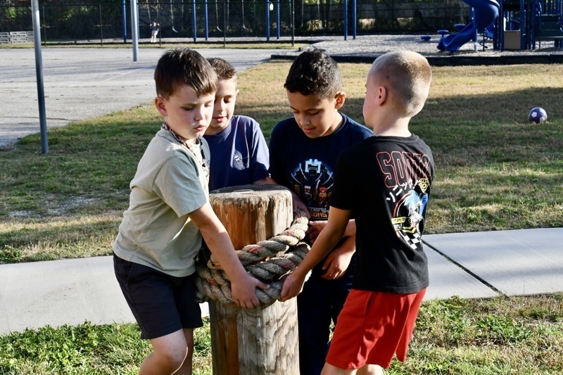 Four people are wrapped around a wooden post with a thick rope on a playground.