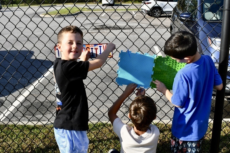 Three people attach colorful mats to a chain-link fence in a parking lot.