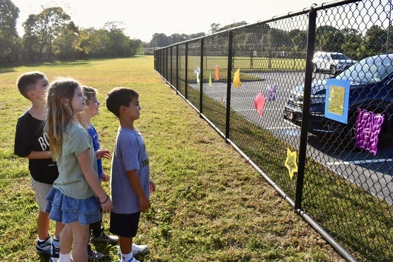 A group of children observes colorful decorations hanging on a fence in a grassy area.