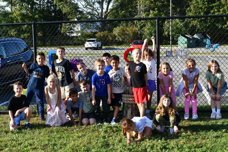 A group of children poses together in front of a fence and parked cars.