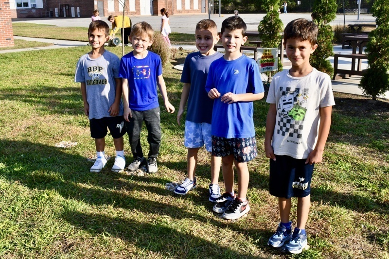 A group of children stands together on grass, wearing casual clothes and sneakers.