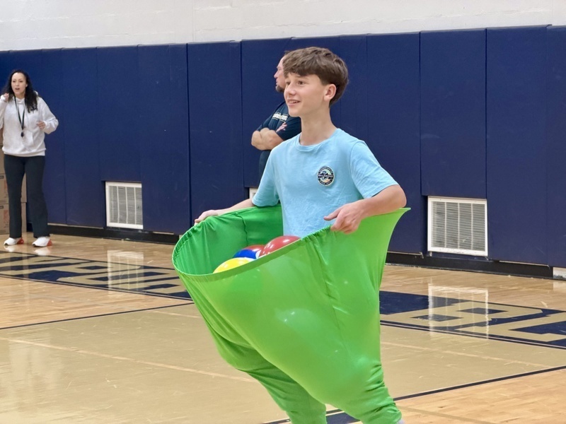A person wearing a green sack holds colorful balls in a gymnasium setting.