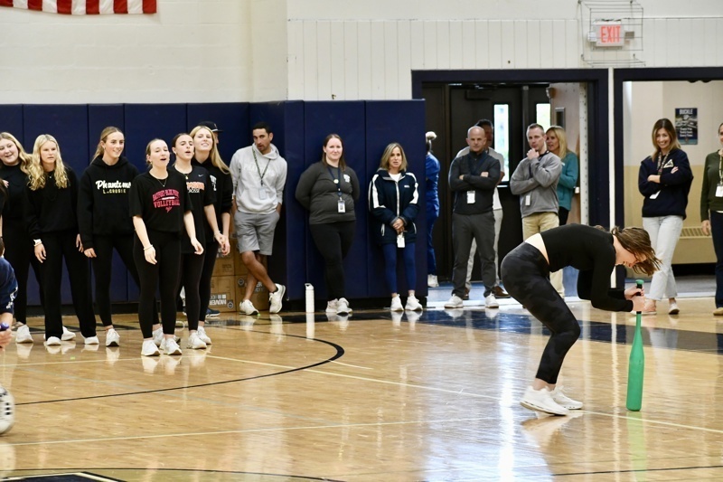 A group watches as a person prepares to swing a green object on a gym floor.