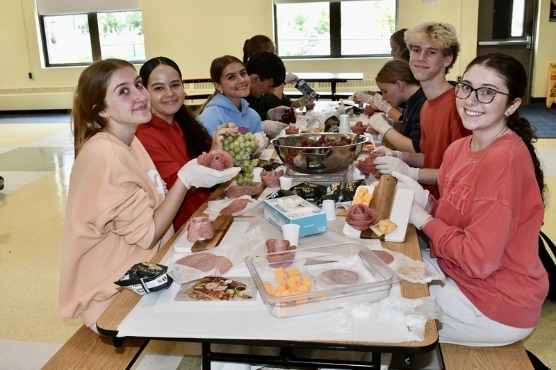 A group prepares food items at a table with a variety of meats and cheeses.
