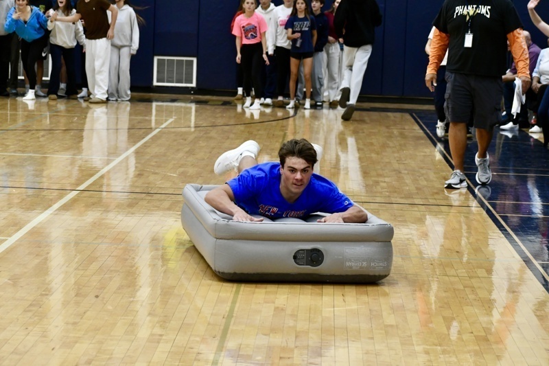 A person slides on an inflatable mattress while others watch in a gymnasium setting.