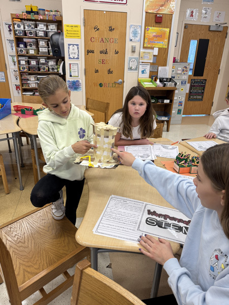 Children engaged in a classroom activity, working collaboratively with craft materials like popsicle sticks and masking tape. The atmosphere is focused and creative.