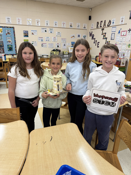 Children engaged in a classroom activity, working collaboratively with craft materials like popsicle sticks and masking tape. The atmosphere is focused and creative.