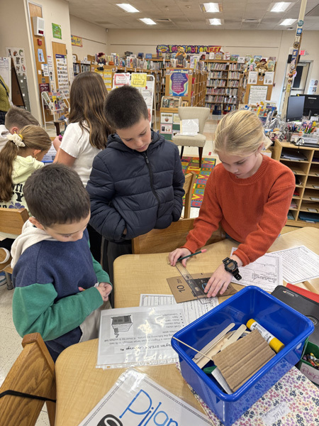 Children engaged in a classroom activity, working collaboratively with craft materials like popsicle sticks and masking tape. The atmosphere is focused and creative.