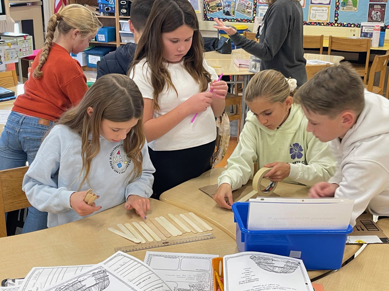 Children engaged in a classroom activity, working collaboratively with craft materials like popsicle sticks and masking tape. The atmosphere is focused and creative.