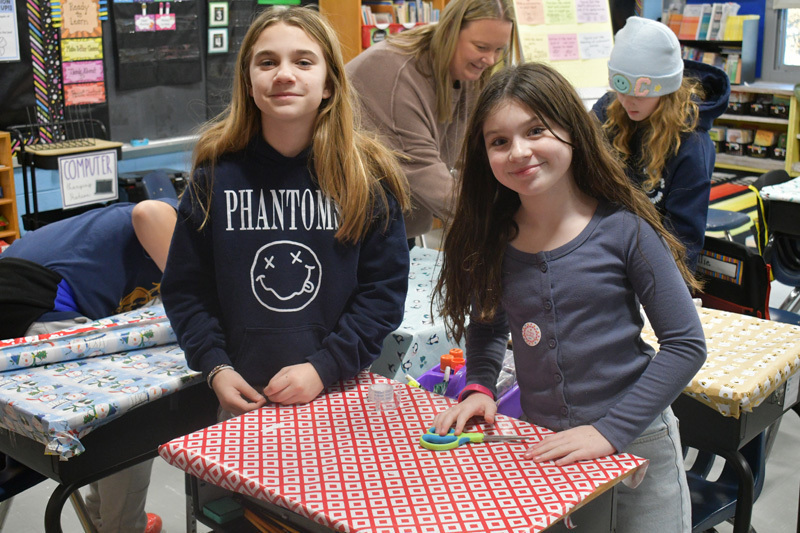 smiling students decorate classroom desks with festive holiday wrapping paper.