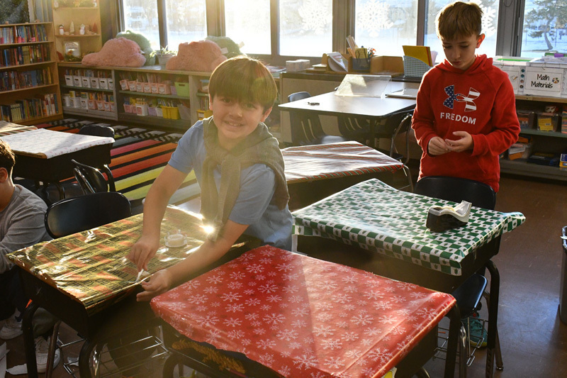 smiling students decorate classroom desks with festive holiday wrapping paper.