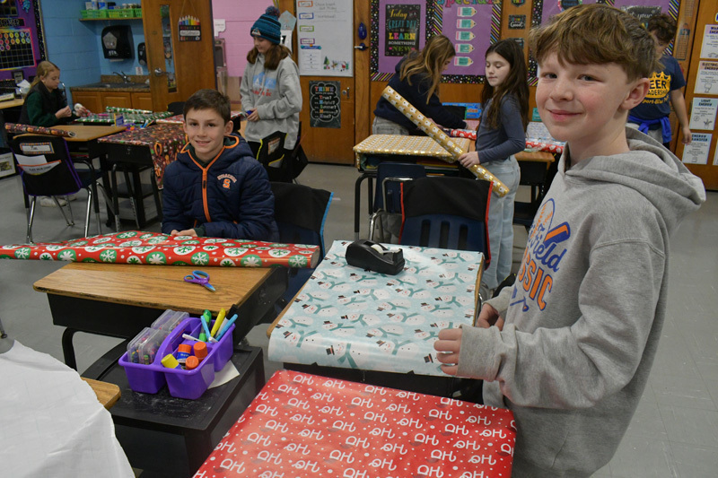 smiling students decorate classroom desks with festive holiday wrapping paper.