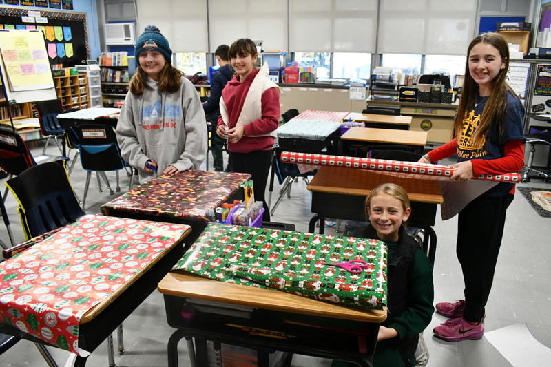 smiling students decorate classroom desks with festive holiday wrapping paper.