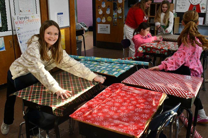smiling students decorate classroom desks with festive holiday wrapping paper.