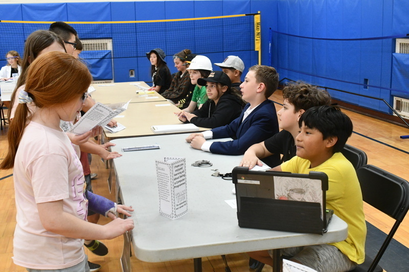Students showcase their projects at a school science fair, featuring creative displays on various topics, including filmmaking and hobbies, in a lively gymnasium setting.