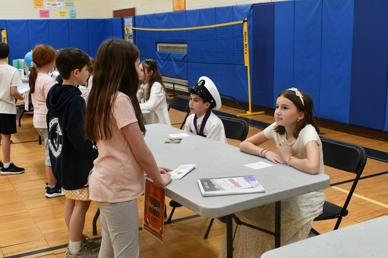 Young students engaged in a classroom activity, interacting at tables with props and costumes, fostering creativity and learning in a vibrant gymnasium setting.