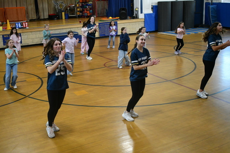 A group of young dancers in school uniforms perform a dance routine on a gymnasium floor, showcasing energetic movements and teamwork.
