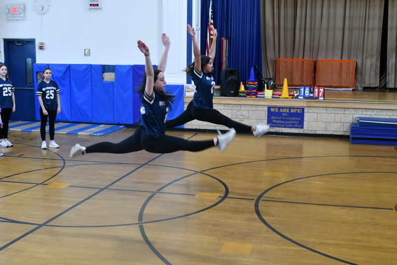 A group of young dancers in school uniforms perform a dance routine on a gymnasium floor, showcasing energetic movements and teamwork.