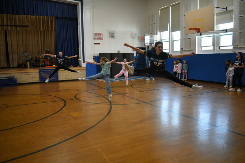 A group of young dancers in school uniforms perform a dance routine on a gymnasium floor, showcasing energetic movements and teamwork.