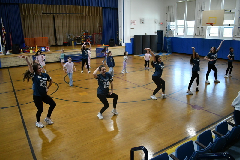 A group of young dancers in school uniforms perform a dance routine on a gymnasium floor, showcasing energetic movements and teamwork.