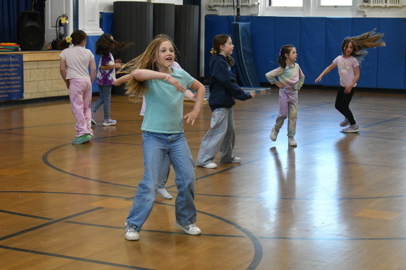 A group of young dancers in school uniforms perform a dance routine on a gymnasium floor, showcasing energetic movements and teamwork.