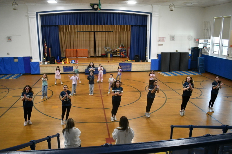 A group of young dancers in school uniforms perform a dance routine on a gymnasium floor, showcasing energetic movements and teamwork.