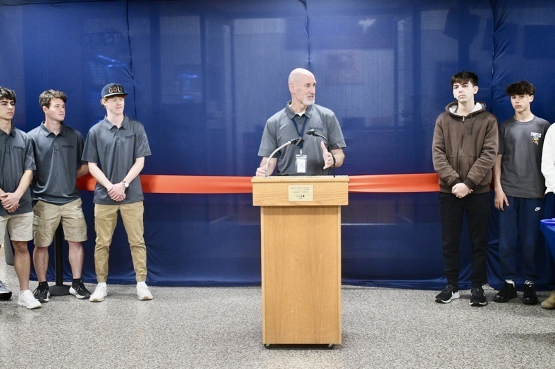 Community event featuring a speaker at a podium with students in matching shirts, celebrating a significant milestone with a red ribbon in the background.