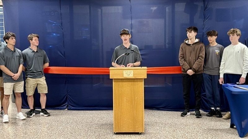 Community event featuring a speaker at a podium with students in matching shirts, celebrating a significant milestone with a red ribbon in the background.