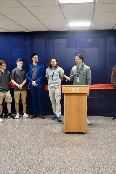 Community event featuring a speaker at a podium with students in matching shirts, celebrating a significant milestone with a red ribbon in the background.