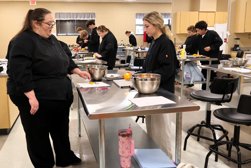 The Culinary Arts teacher is working with a student on making garnishes while other students are in the background working on theirs. 