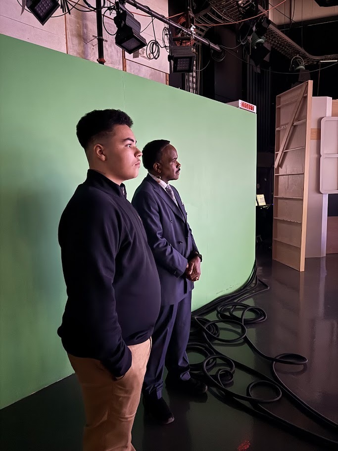 Student stands with meterologist in front of green screen.